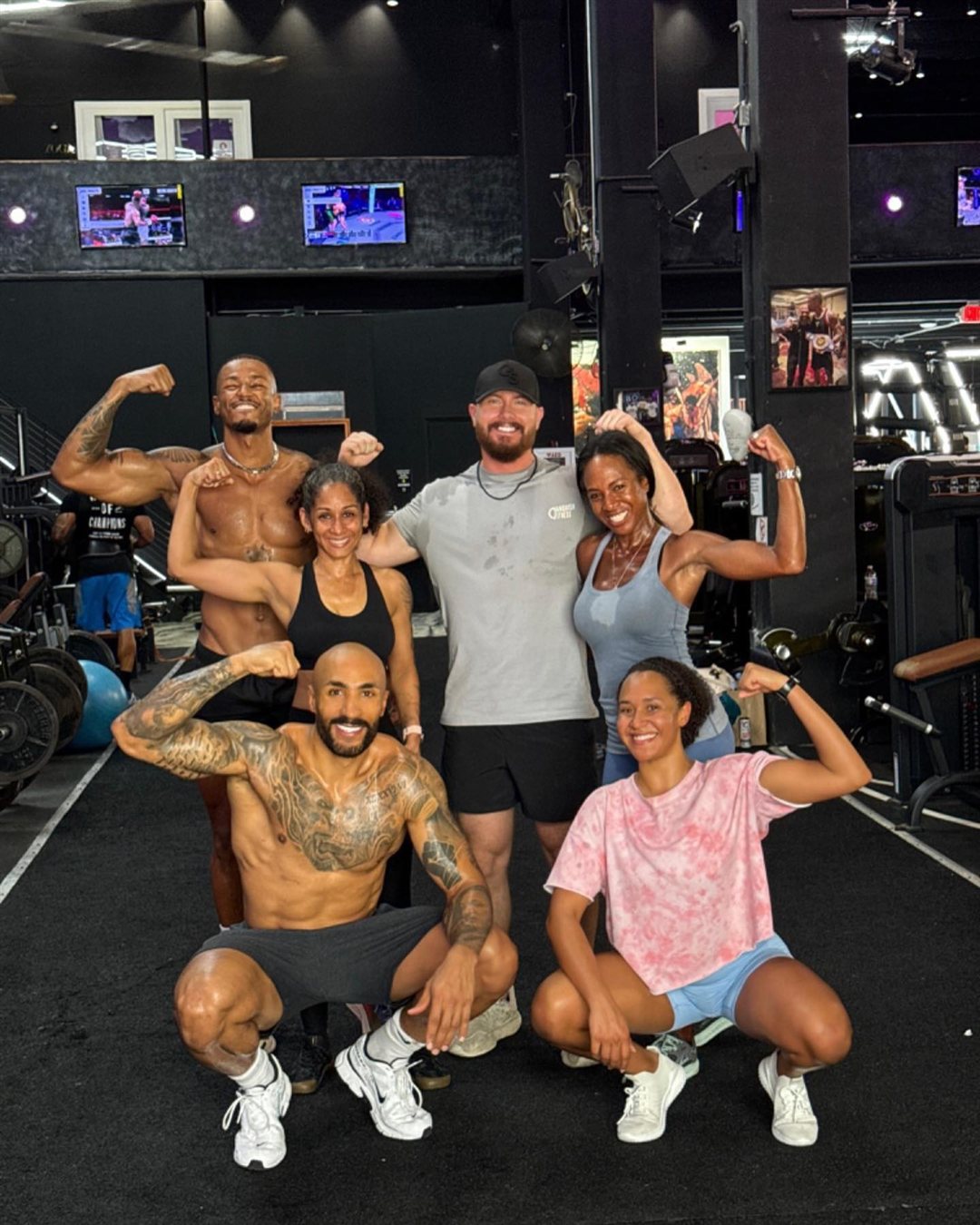 Jot with a group of his clients in the gym, everyone flexing and smiling.